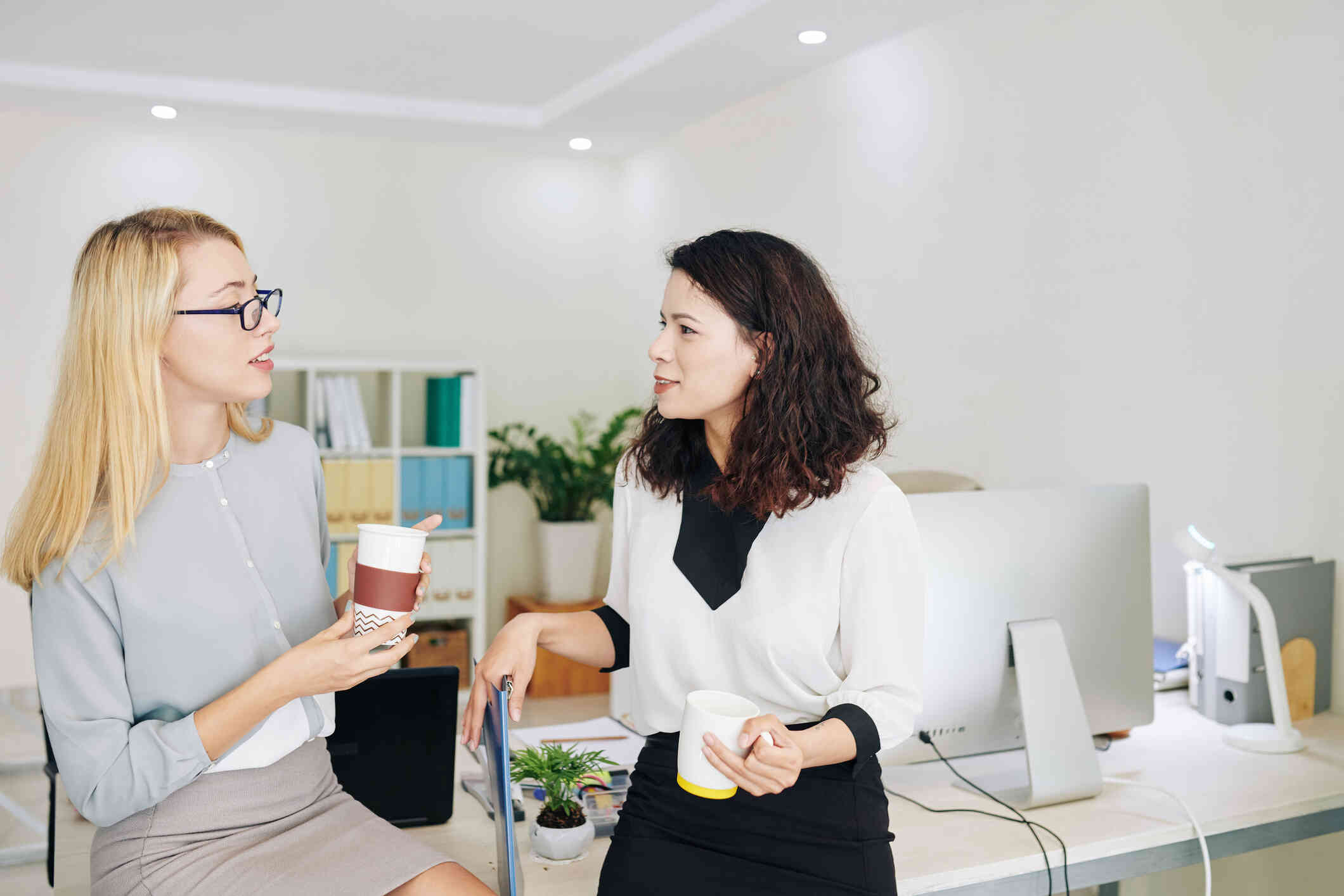 Two female coworkers stand side by side in the office and talk while holdings cups of coffee.